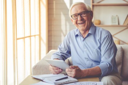 Handsome old man in eyeglasses is using a digital tablet, looking at camera and smiling while working at home